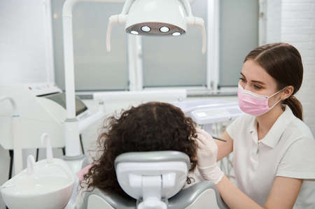 Young female dentist hygienist in protective medical mask and gloves performing oral treatment in contemporary dental clinic. Dentistry, oral care and dental hygiene conceptの写真素材