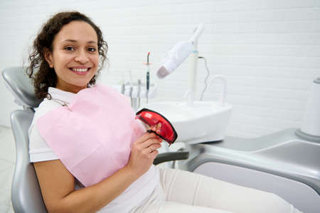 Smiling woman with beautiful toothy smile sits in dentists chair and holds UV protective goggles...