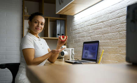 Confident female doctor holds a dental impression of human jaw looking at camera sitting in front of...