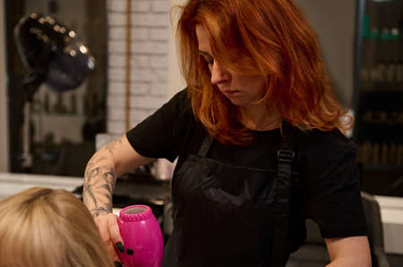 Close-up. Middle-aged red-haired female hairdresser with tattooed arms in a strict black uniform combing, straightening and drying the blonde hair of a client of a beauty salon using a pink hairdryerの写真素材