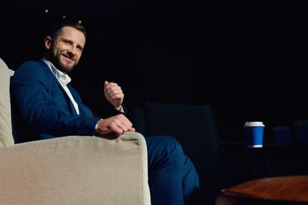 Handsome attractive young Caucasian man in business suit sits in an armchair in the lobby of a luxury hotel, enjoys a coffee break during a business trip and looks confidently to the side cute smilingの写真素材