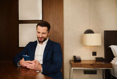 Confident portrait of a handsome prosperous business traveler, successful businessman, entrepreneur, investor in dark suit resting in hotel room, smiles while using mobile phone during business tripの写真素材