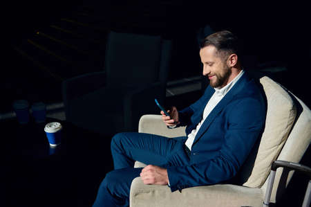 Handsome, confident successful businessman sits in an armchair near a table with a cup of coffee in...