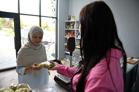 Cheerful Arab Muslim woman with covered head in hijab holding sealed olives while making purchases in a groceryの写真素材