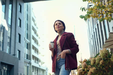 Self-confident beautiful young woman with takeaway paper cup of coffee on the urban background of...