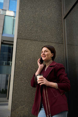Cheerful young European woman in casual wear talks on mobile phone standing by the marble wall of a...