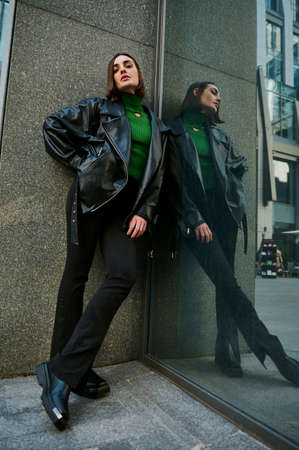 Full-length fashion portrait of an attractive European woman in eco-leather clothes looking at camera, leaning against the marble wall of a modern building reflected on the glass entranceの写真素材