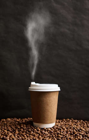 Close-up of a takeaway cardboard cup of hot drink with steam on a surface with scattered roasted coffee beans on a black wall background with copy space for advertisementの写真素材