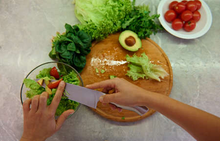 High angle view of female chef hands with a kitchen knife slicing tomatoes and throwing them into a glass transparent bow while preparing healthy raw vegan saladの写真素材