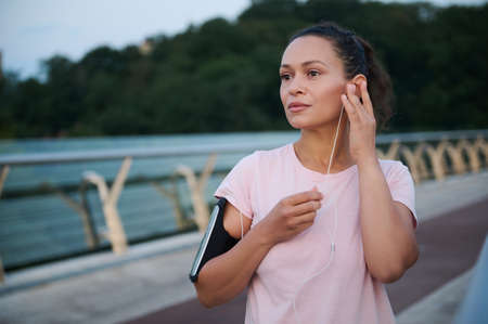 Determined young athlete pretty woman puts on earphones while standing on the bridge before morning...