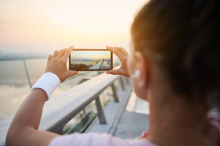 Rear view of athlete woman jogger in pink t-shirt holding smartphone and taking photo of beautiful sunrise at the end of the bridge before morning jog. Mobile phone in live view mode. Outdoor workoutの写真素材