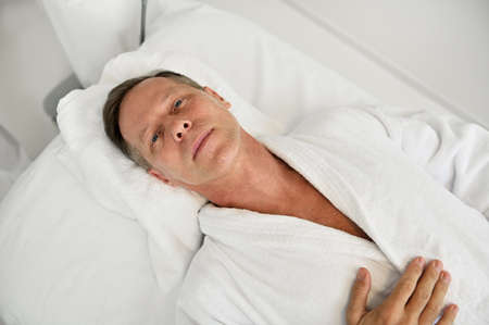 High angle view of handsome relaxed mature 50 years old European man in white terry bathrobe looking at camera while lying on massage table in white luxury wellness spa clinicの写真素材