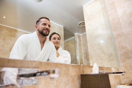 Charming young couple in love, newlyweds on their honeymoon enjoying togetherness, embracing together standing in the bathroom, looking into their mirror reflection. Love, affection, tenderness.の写真素材