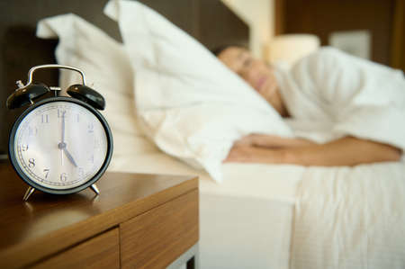 Soft focus on black alarm clock on nightstand next to a blurred bed with sleeping woman. Copy space...