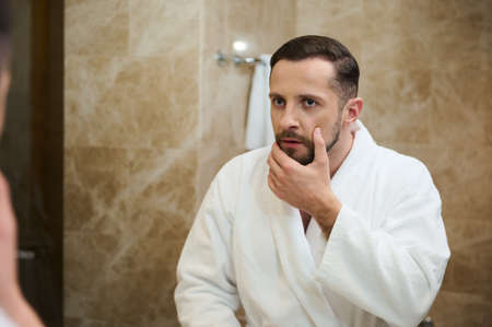 Handsome young European man in a white terry bathrobe touching the stubble before shaving, looking in surprise at his mirror reflection in the bathroomの写真素材
