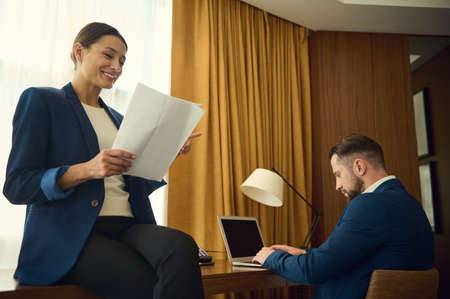 Beautiful smiling middle aged successful delighted woman in casual suit sitting on a table with documents while her partner by business, typing on laptop keyboard working together in hotel roomの写真素材
