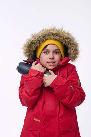 School-age traveler adventurer handsome boy warming himself hugging thermos, shivering from cold looking at camera isolated on white background. Copy space for ads. Winter leisure, holidays conceptの写真素材