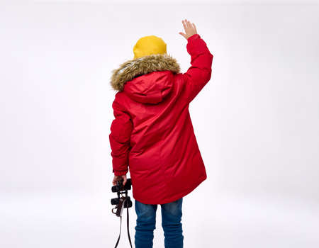 Rear view of elementary aged boy in bright down jacket with hood and a woolen hat holding binoculars and waving hello with his raised up hand. Copy space for ads. Tourism and winter travel conceptの写真素材