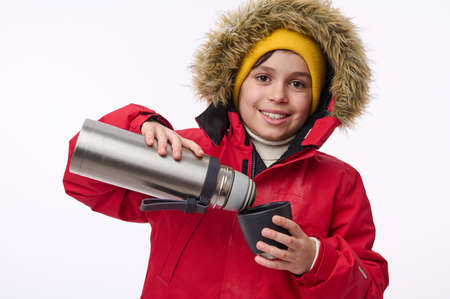 Horizontal portrait of adorable school-age boy winter traveler adventurer pouring hot drink from flask into mug smiling toothy smile looking at camera isolated on white background with copy spaceの写真素材