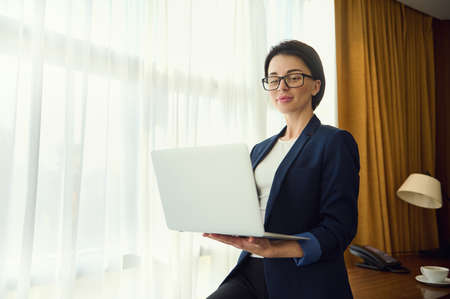 Prosperous concentrated woman, entrepreneur, journalist, sales manager in business casual suit working on laptop standing by window in hotel room. Cope space for advertisement textの写真素材