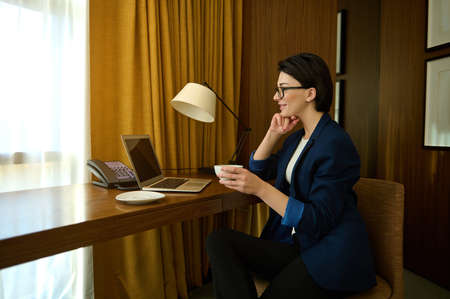 Beautiful young prosperous businessman, successful female sales manager focused on writing a progress report on a laptop, sitting at the table and drinking coffee in her officeの写真素材