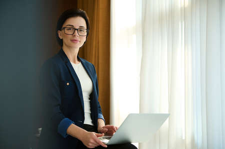 Business portrait of a beautiful self-confident young businesswoman, successful sales manager, investor, female investment advisor wearing eyeglasses, holding laptop and confidently looking at cameraの写真素材