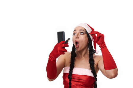 Beautiful European woman in Santa carnival costume, looking at her reflection on the smartphone screen, putting mascara on her eyelashes. Final makeup touches before celebrating the Christmas holidaysの写真素材