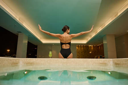 Rear view of middle aged European woman with beautiful aesthetic slim body in black swimsuit looking to the side sitting on edge of thermal bathtub with hydromassage therapy in luxury spa resortの写真素材