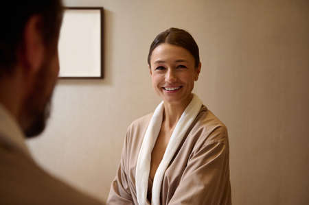 Delighted middle aged European woman in bathrobe smiling looking at her blurred husband on the foreground, resting at wellness spa resort on honeymoon. Cute couple getting beauty treatment togetherの写真素材