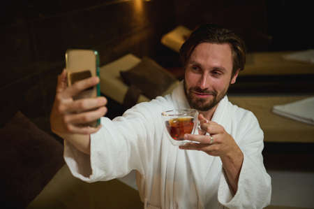 Handsome Caucasian man, sitting by bathtub in lounge area, drinking tea and looking at camera on smartphone in his outstretched hand, making selfie, enjoying holidays in luxurious wellness spa resortの写真素材