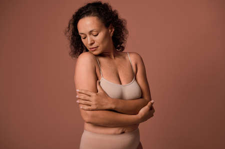 Confident serene gorgeous middle aged woman with imperfect body, cellulite and stretch marks after childbirth, in beige underwear, looking down, hugging herself on colored background with copy spaceの写真素材
