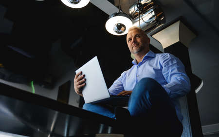 View from the bottom to a successful middle aged European man in casual denim jeans and blue shirt working on laptop sitting on a table at home kitchen, working remotely from homeの写真素材
