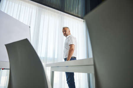 Hnadsome middle aged entrepreneur, successful sales manager, businessman standing near flip chart with copy space for advertisement text, and confidently looking at cameraの写真素材