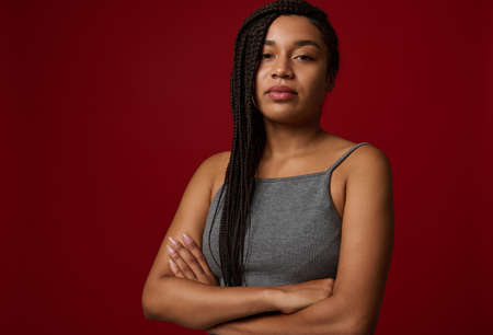 Waist length portrait of a young African woman with dreadlocks in gray sports t-shirt confidently looks at camera, posing with crossed arms against red colored background with copy spaceの写真素材