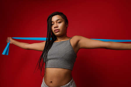 Attractive young African sportswoman wearing wireless headphones, stretching elastic band during her exercise routine, working out against red colored background with space for adの写真素材