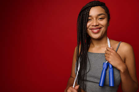 Cheerful African woman with dreadlocks posing against a red background with a skipping rope around her neck, smiling with a toothy smile, looking at the cameraの写真素材