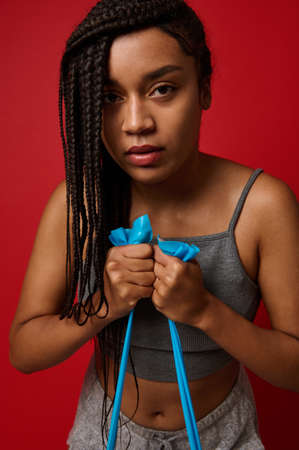 Close-up portrait of a strong African woman, female athlete with stylish dreadlocks, exercising with resistance band against red colored background with copy space for advertisementの写真素材