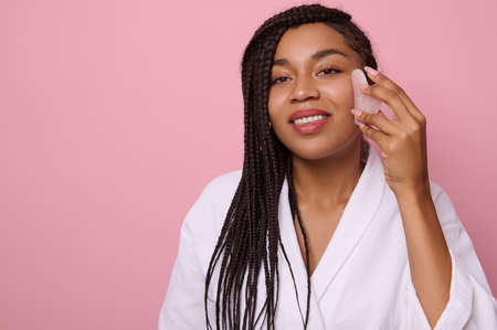 Beautiful African woman with dreadlocks, wearing white waffle bathrobe, smiles toothy smile looking at camera while doing facial smoothing massage with jade Gua-Sha massager, against pink backgroundの写真素材