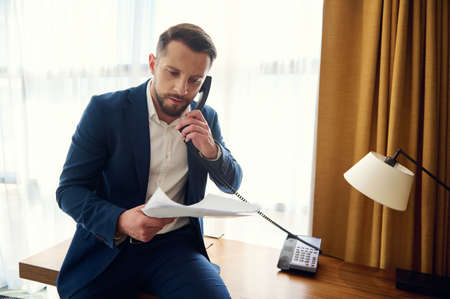 Busy puzzled handsome middle aged European man, entrepreneur, successful businessman holding a stack of documents and negotiating while talking on landline phone in hotel room during business travelの写真素材