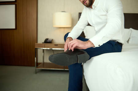 Focus on the shoes of a young bearded Caucasian businessman. Handsome middle aged man taking off shoes sitting on edge of bed in hotel room relaxing after business meetingsの写真素材