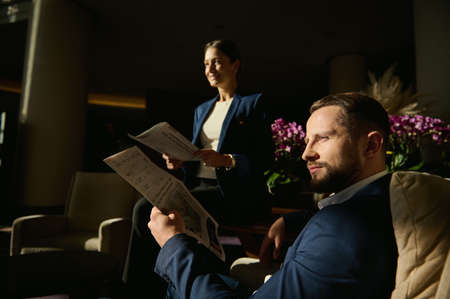 Handsome business partners, colleague, team, middle aged Caucasian man and woman sitting on armchair in the hotel lobby and reading newspaper looking away. Business, people, trip conceptの写真素材