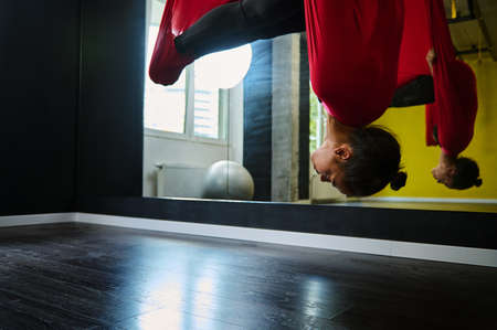 Young beautiful African woman with slim body hanging on a red hammock, practicing aerial fly yoga stretching exercises with anti-gravityの写真素材