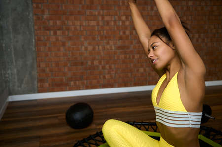African sportswoman in bright yellow tracksuit raising arms up, stretching her body, sitting in...