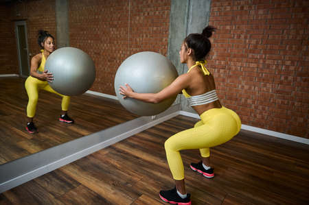 Rear view of a sporty woman holding a gray fitness ball and doing glute exercises, pumping buttocks...