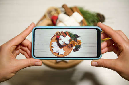 Phone in real time. Hands holding phone and taking pictures of gingerbreads laid out on wooden board and on baking paper sprinkled with sugar powder next to dried orange slices, pine cones and twigsの写真素材