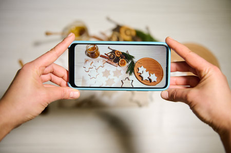 Phone in real time. Hands holding phone and taking pictures of gingerbreads laid out on wooden board and on baking paper sprinkled with sugar powder next to dried orange slices, pine cones and twigsの写真素材
