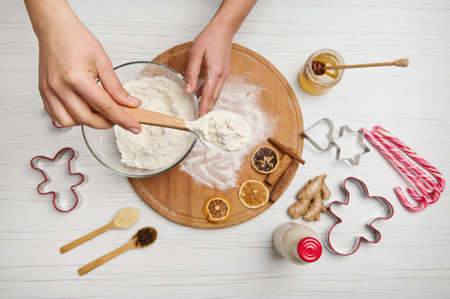 Flat lay of female hands holding a spoon with flour above a bowl on a wooden table with dried orange...