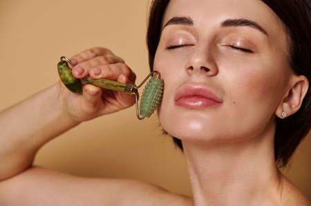 Young Caucasian brunette doing anti-aging, smoothing, lymphatic drainage facial massage with jade roller massager. Skin care. Attractive natural beauty woman massages her face with stone roller.の写真素材
