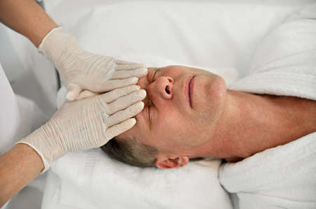 Close-up of a mature European man relaxing during face lifting massage while a beautician...