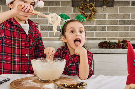 Charming Caucasian children cooking Christmas cookies together at home kitchen. Adorable boy kneads dough with a wooden spoon when his cute younger sister climbs her hands into the dough and tastes itの写真素材